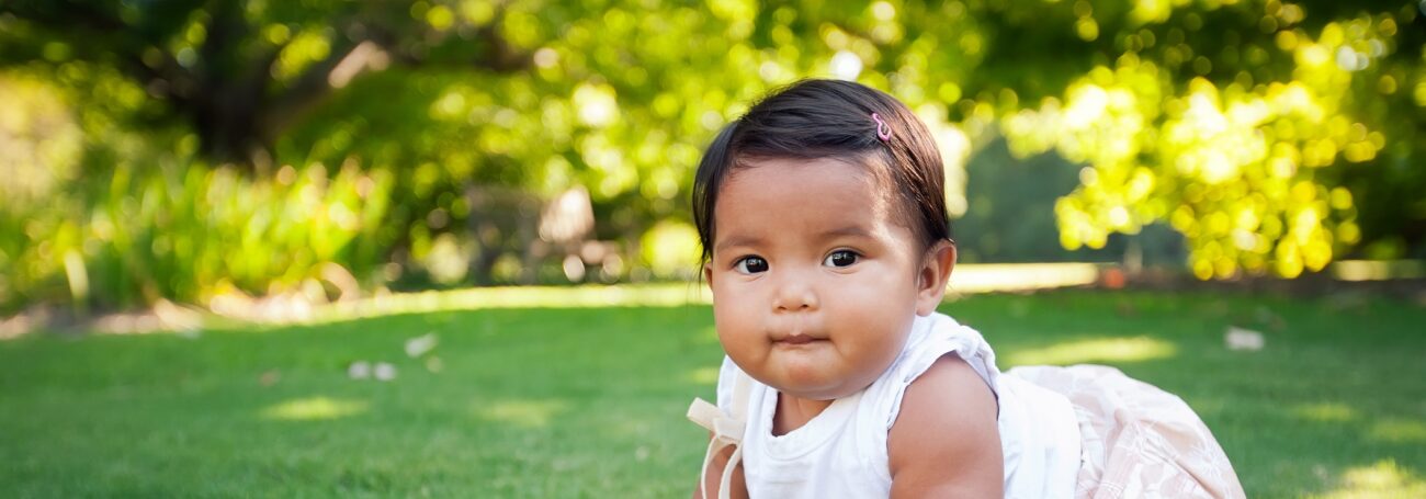 Toddler girl with brown hair and eyes stares into camera while crawling outside