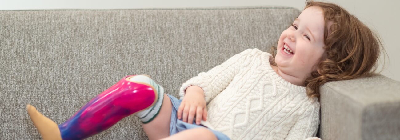 Young girl with prosthetic right leg smiles and lounges on a tan couch