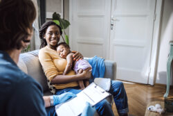 Woman cradles baby while sitting on her couch and talking to nurse home visitor