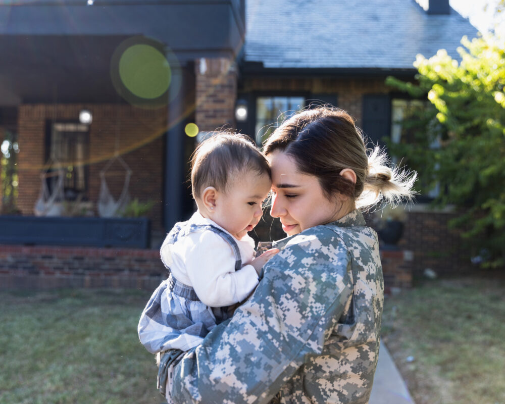 Woman wearing Army fatigues hold infant outside of their home