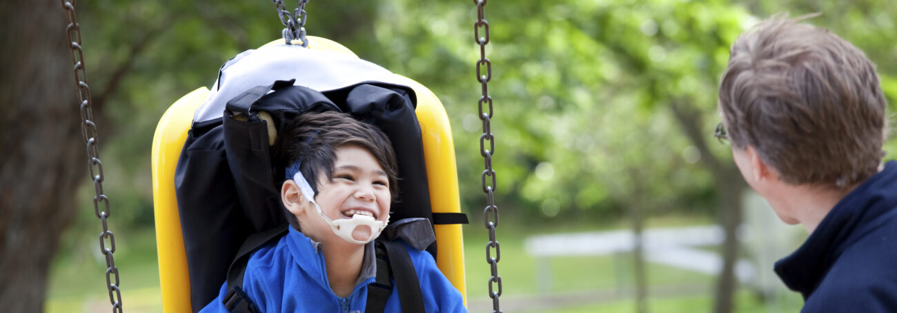 A young boy with a disability smiles in a handicap-accessible swing while his dad looks on