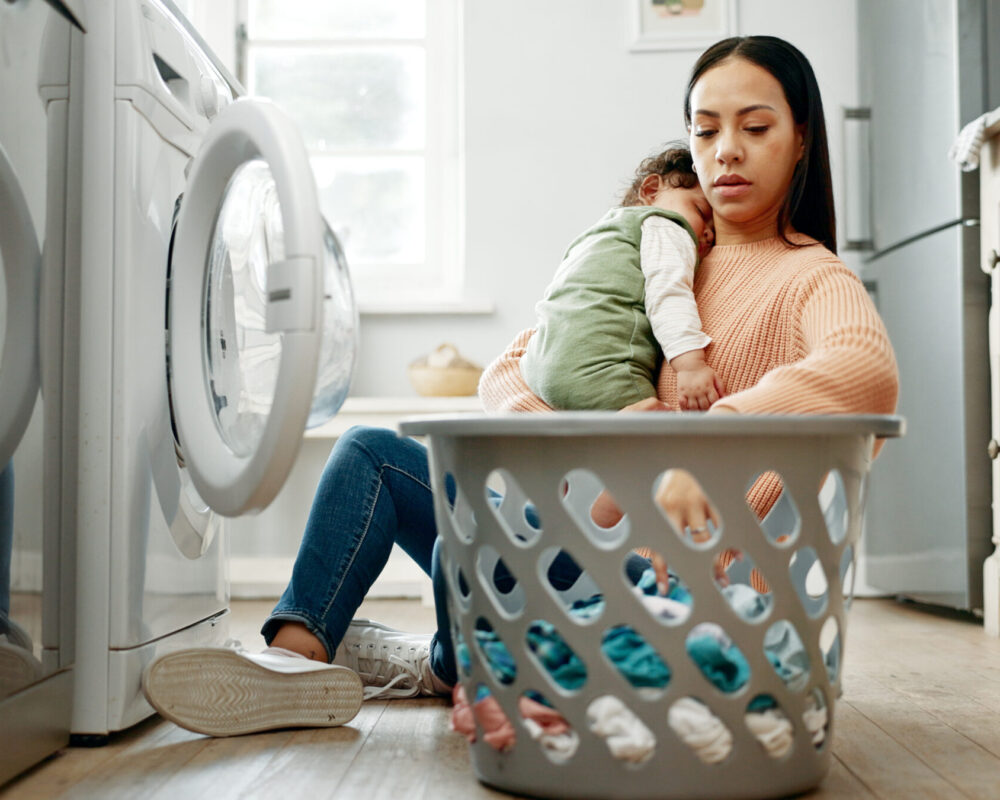 Mother holds sleeping infant while doing laundry