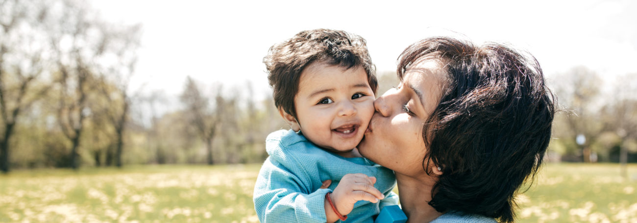 Mother kissing toddler daughter dressed in blue on the check while standing outside