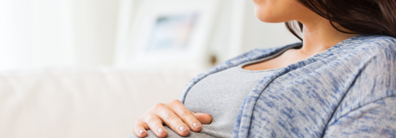 Woman in gray shirt cradles her pregnant belly as she looks off to the side