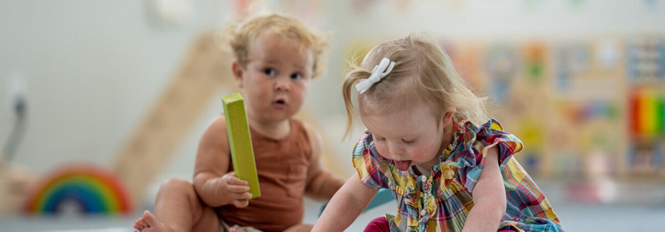 two toddlers sit on the floor and play with blocks