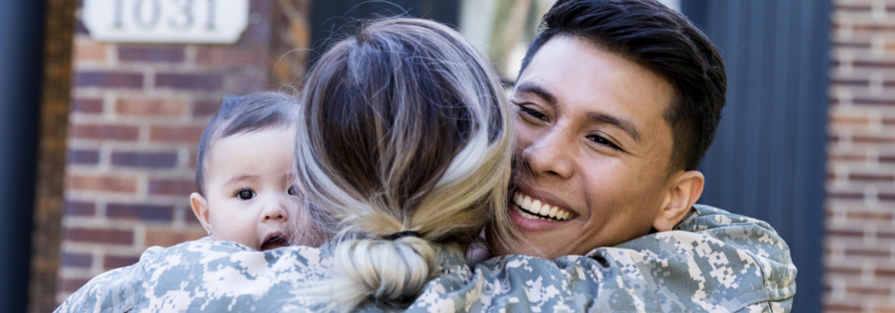 Back of woman in Army uniform as she hugs her husband and baby in front of their house