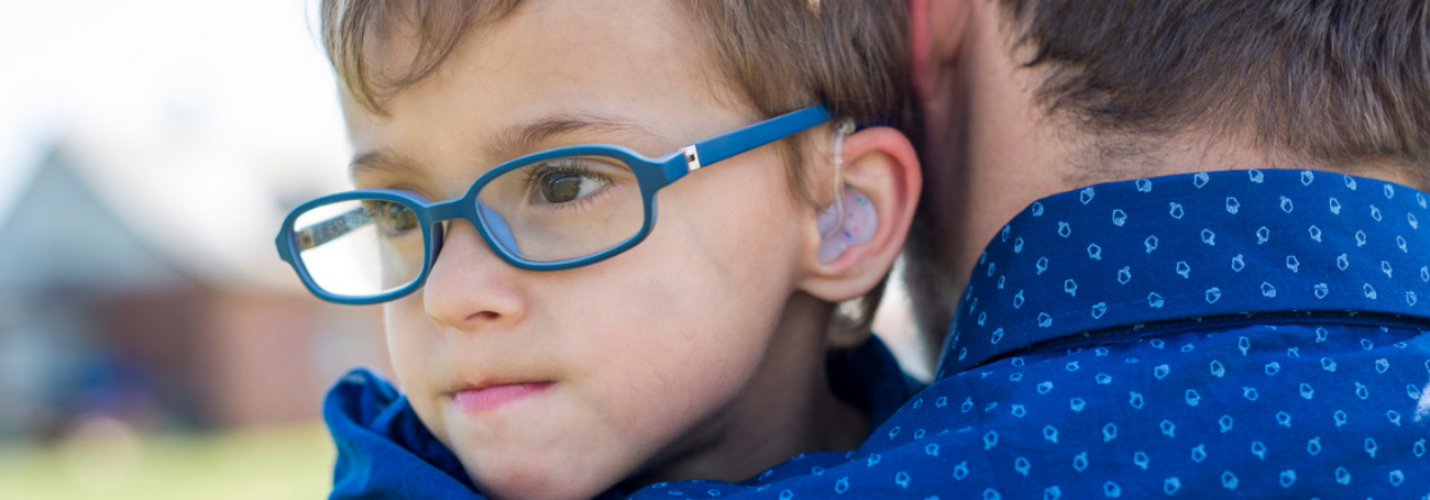 Young boy with blue glasses and hearing aid being carried by his father