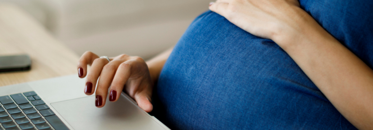 Pregnant woman sits in front of a laptop with her hand resting on her belly